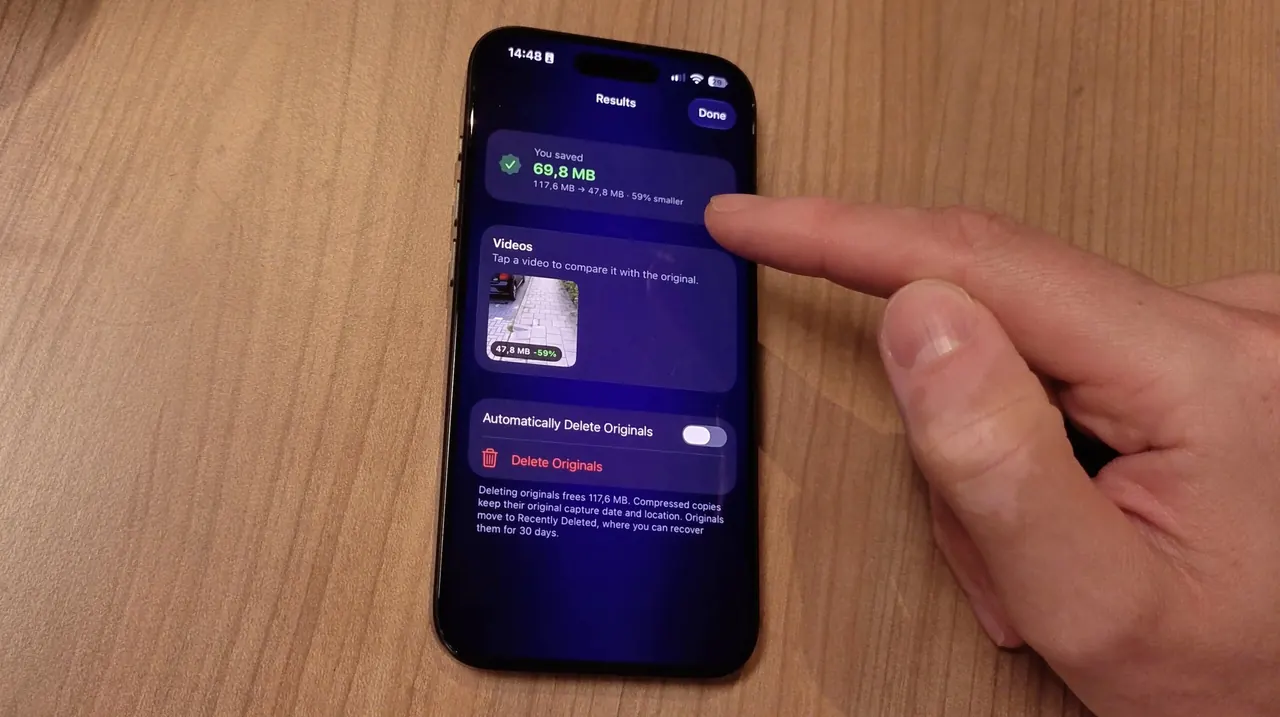 Un iPhone posé sur une table en bois affiche l’écran Results de Video Compressor après la compression d’une vidéo, avec l’interrupteur Automatically Delete Originals désactivé et un doigt qui pointe vers l’écran.