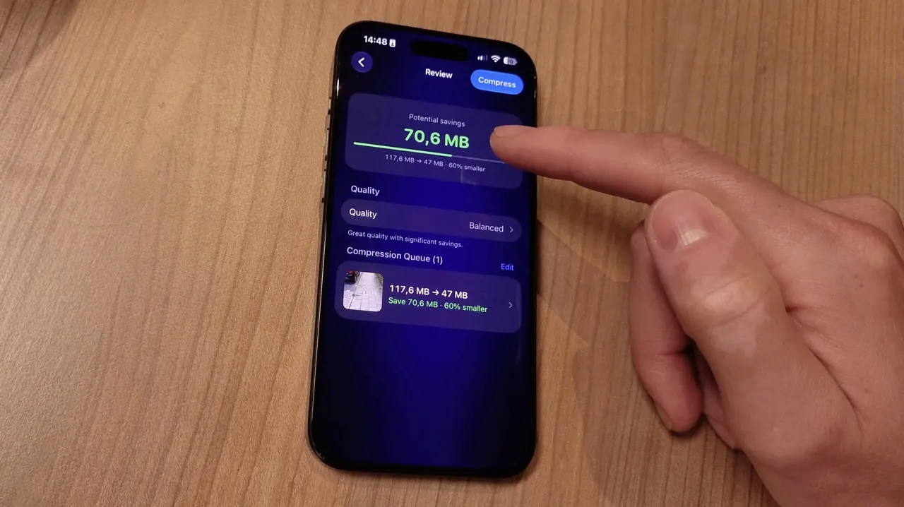 Un iPhone posé sur une table en bois affiche l’écran Review de Video Compressor pour une vidéo, tandis qu’un doigt pointe vers l’écran.